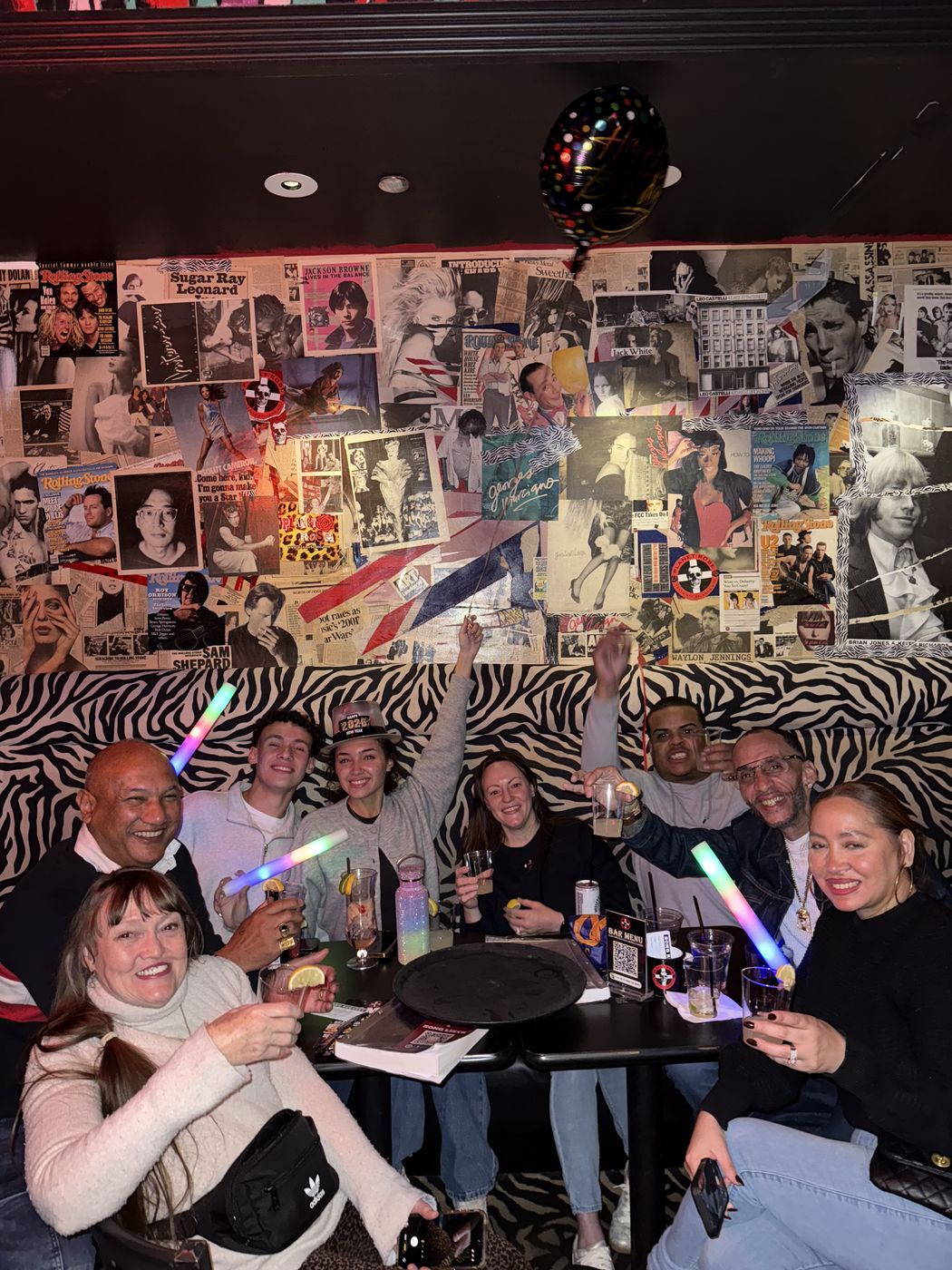 A group celebrating with glow sticks in a booth against the collage wall